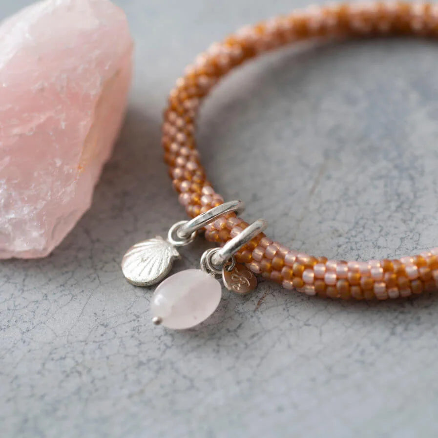 Armband met licht en donker roze rond gevlochten kralen. Er hangt een zilveren bedel van een schelp en een rozenkwarts bedel aan.