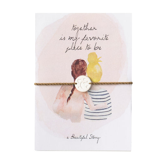 Klein kaartje met waterverf afbeeling van twee vrouwen die elkaar vasthouden en de tekst together is my favorite place to be. Er zit een armbandje bij van lichtbruin waxdraad met een zilverkleurig muntje met hartjes.