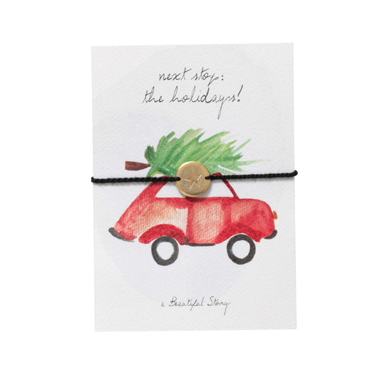 Kerstkaart met armbandje van katoen en goudkleurige muntje met een ster erop. Op het kaartje staat de tekst Next stop the holidays en de waterverf afbeelding is een kleine rode auto met een kerstboom vastgebonden aan het dak.