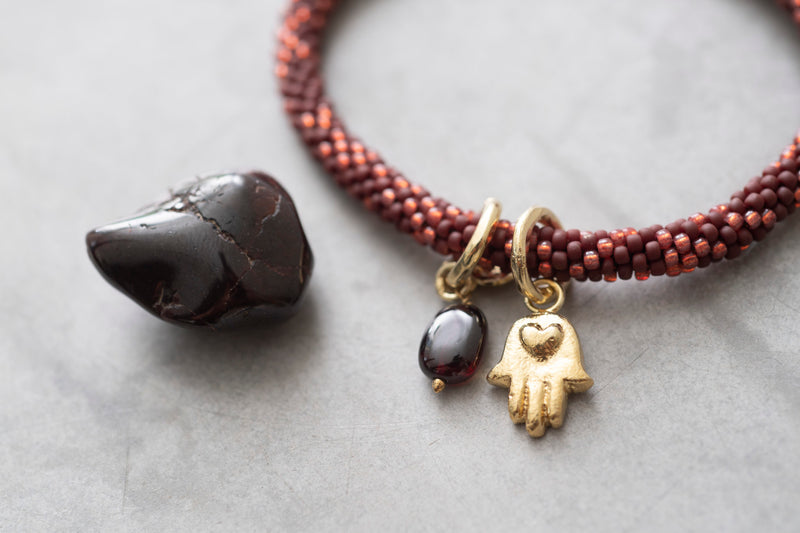 Rond gevlochten armband met lichte en donkerrode glaskralen met een goudkleurige hamsa bedel en granaat steen bedel